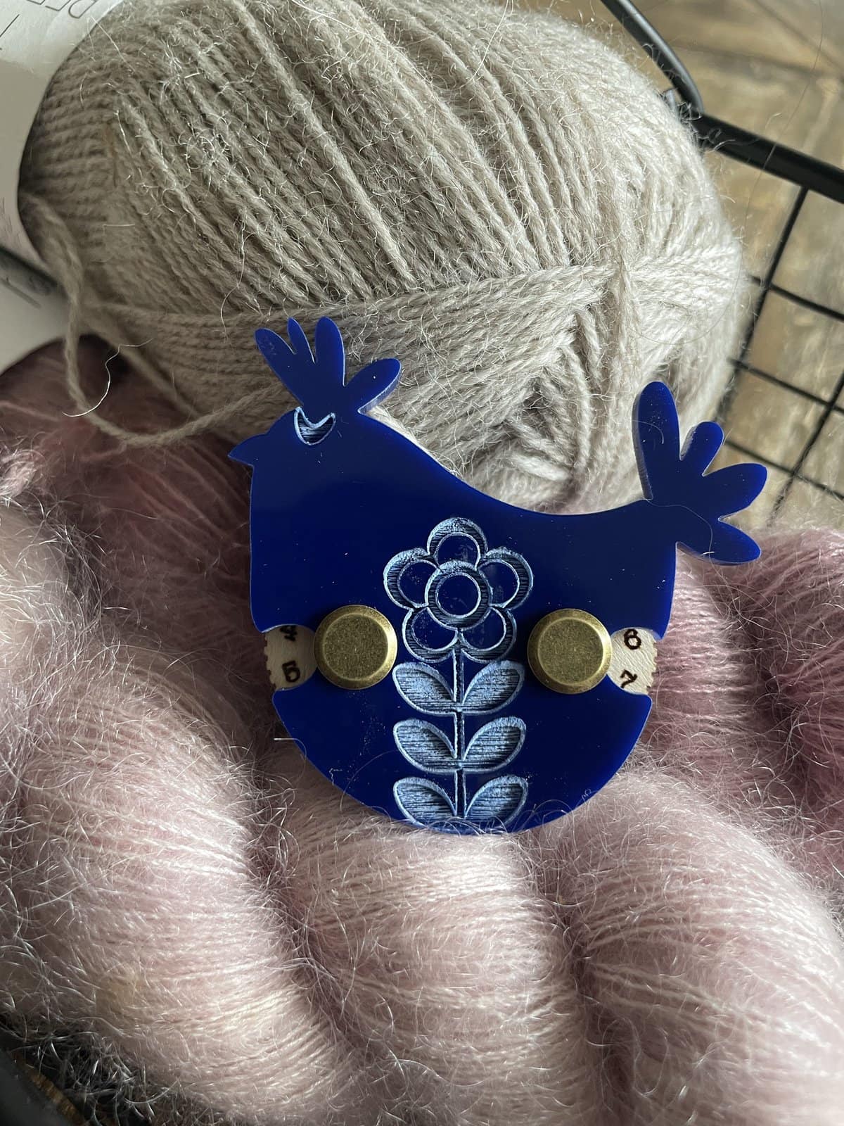 Soft beige yarn ball on a pink fuzzy surface, decorated with a blue moose-shaped pin with floral design, ideal for knitting or crochet projects.