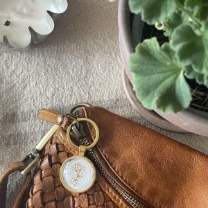 Handwoven brown leather bag with a decorative keychain, potted green plant, and candle on textured surface.