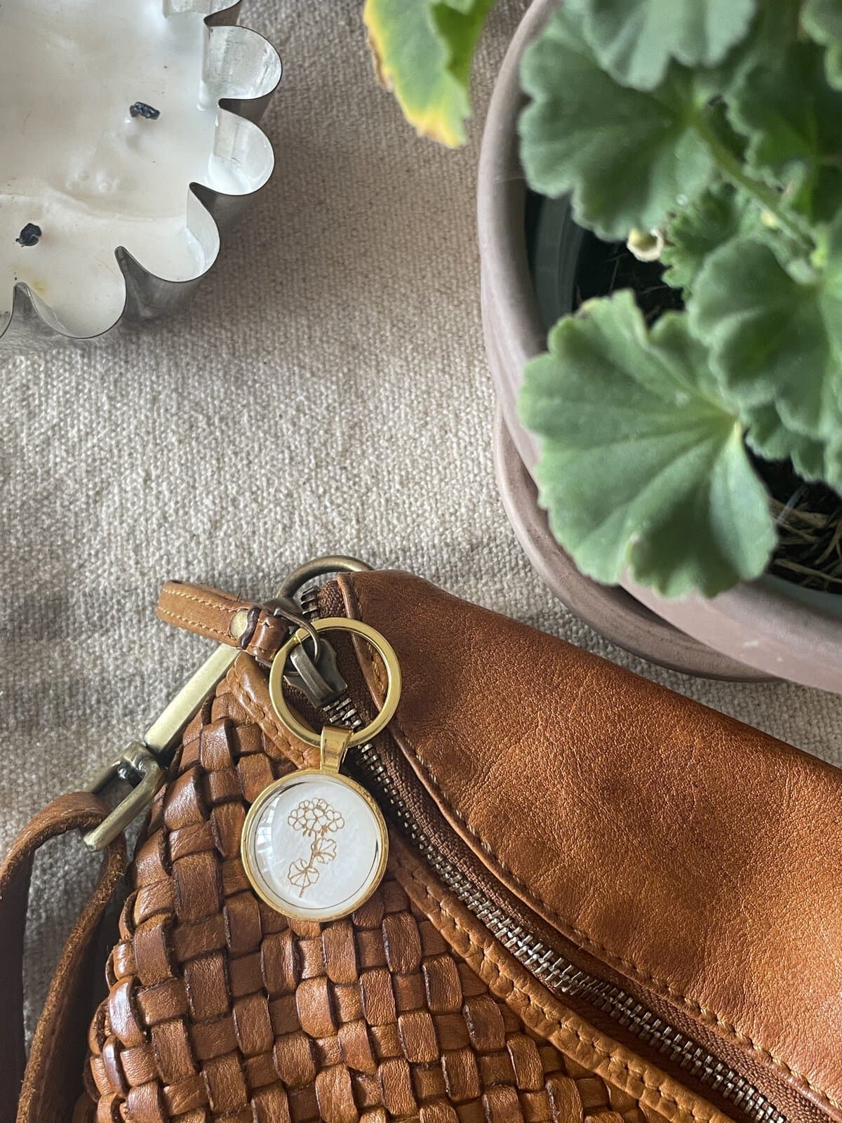 Handwoven brown leather bag with a decorative keychain, potted green plant, and candle on textured surface.