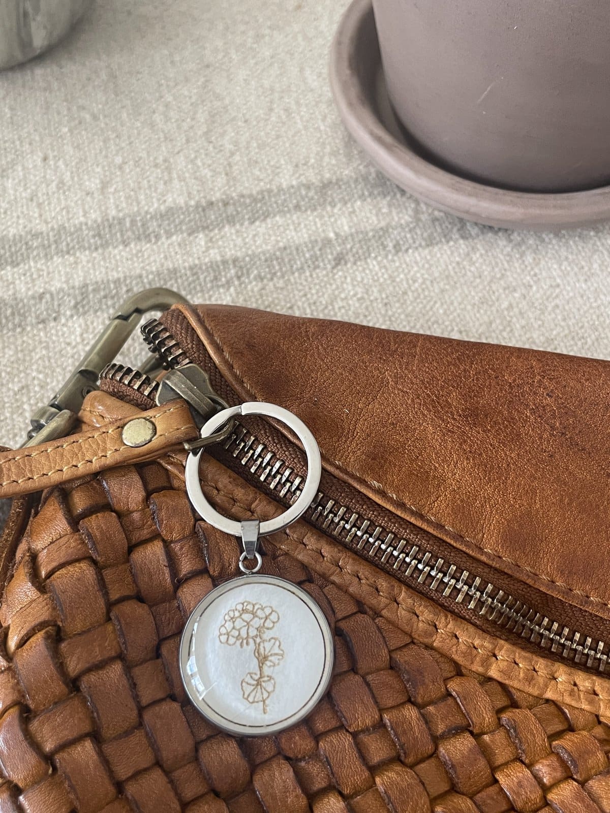 Leather woven handbag with metal hardware and flower keychain on beige fabric surface.