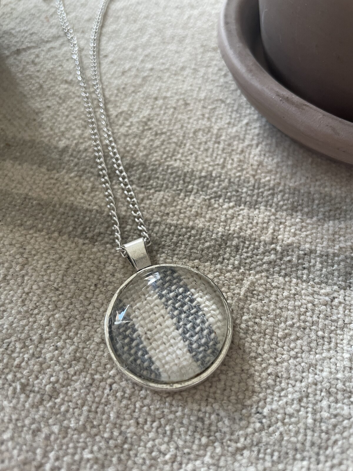 Shimmering silver necklace with circular pendant featuring woven fabric design on a textured beige surface.