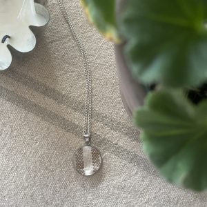 Silver necklace with round pendant and chain on neutral fabric, styled with plant and decorative dish. Perfect for elegant jewelry display.