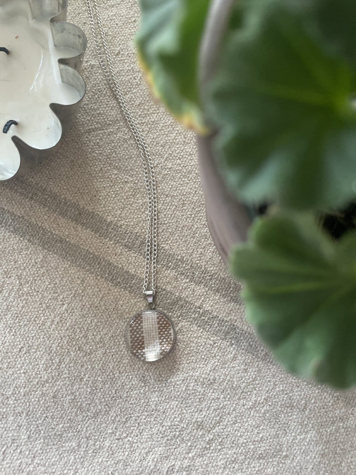 Silver necklace with round pendant and chain on neutral fabric, styled with plant and decorative dish. Perfect for elegant jewelry display.