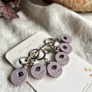 Color-coded stitch marker charms for knitting, labeled 4.5mm and 5.5mm, on a white card with yarn and fabric background.