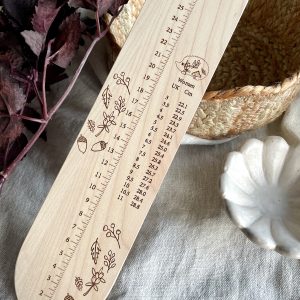 Wooden yarn measurement ruler for knitting and crochet, with decorative botanical illustrations.