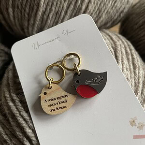 Soft beige yarn with metallic bird-themed enamel charms on Unwrapped Yarn packaging, perfect for knitting or crocheting projects.