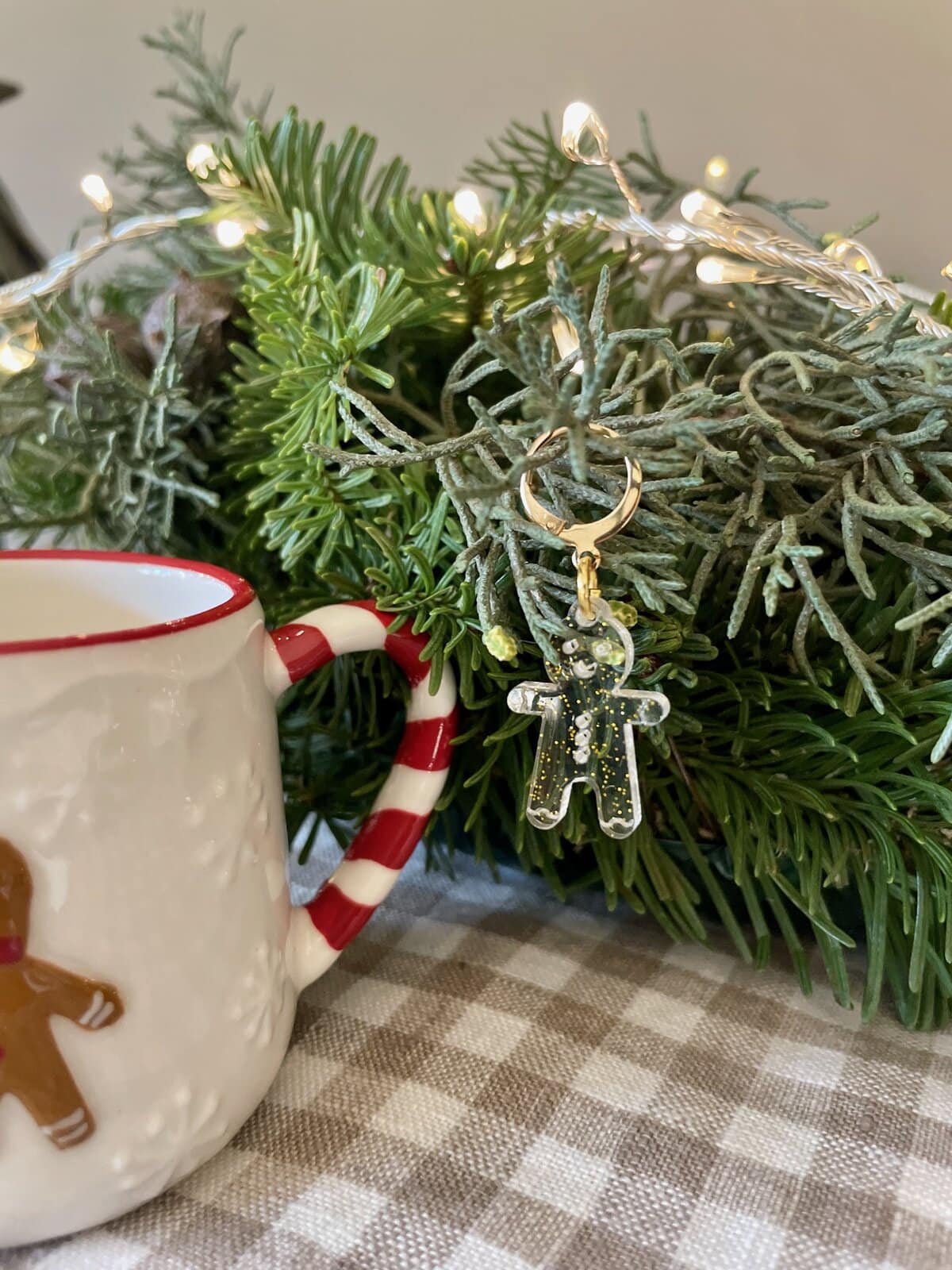 Colorful holiday yarn gingerbread man ornament hanging from festive greenery with a mug nearby.