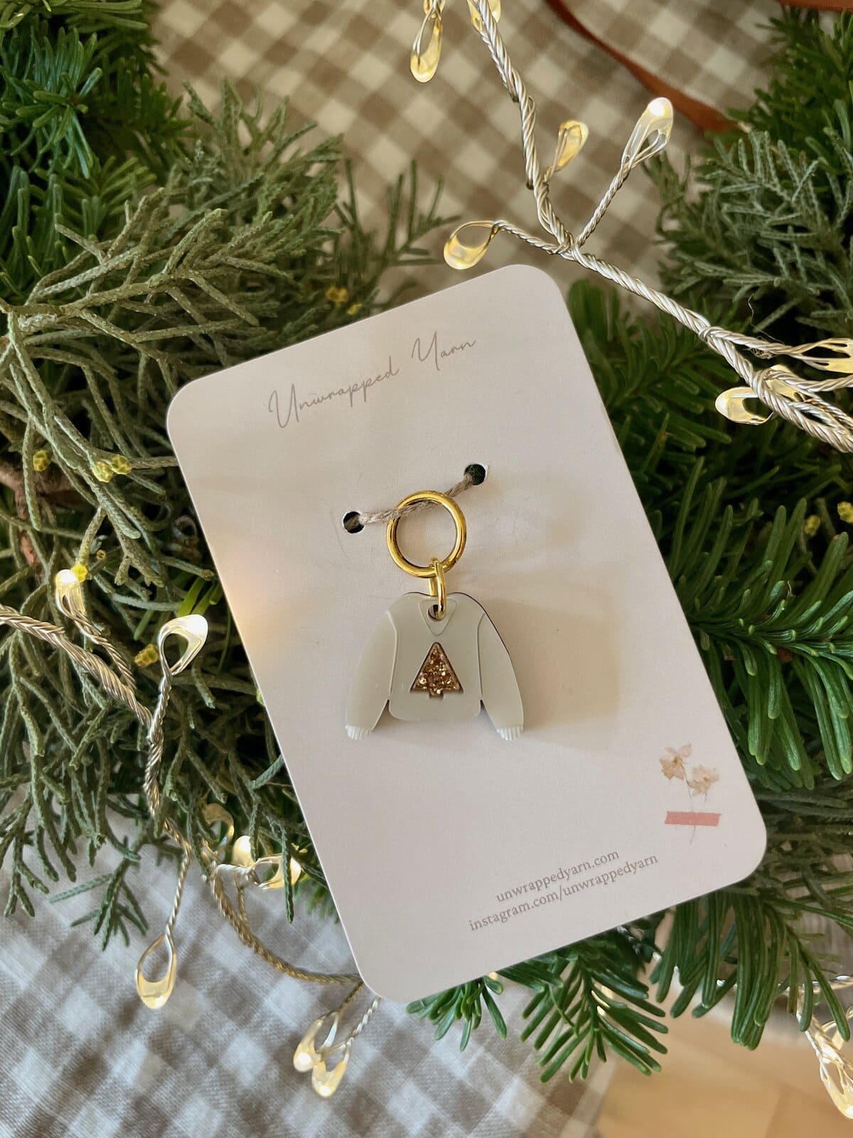 Delicate white sweater jewelry charm with gold arrow detail, gift box, and festive greenery for holiday accessories.