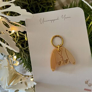 Delicate jewelry charm featuring a sweater design on a white card, surrounded by festive greenery and warm string lights.