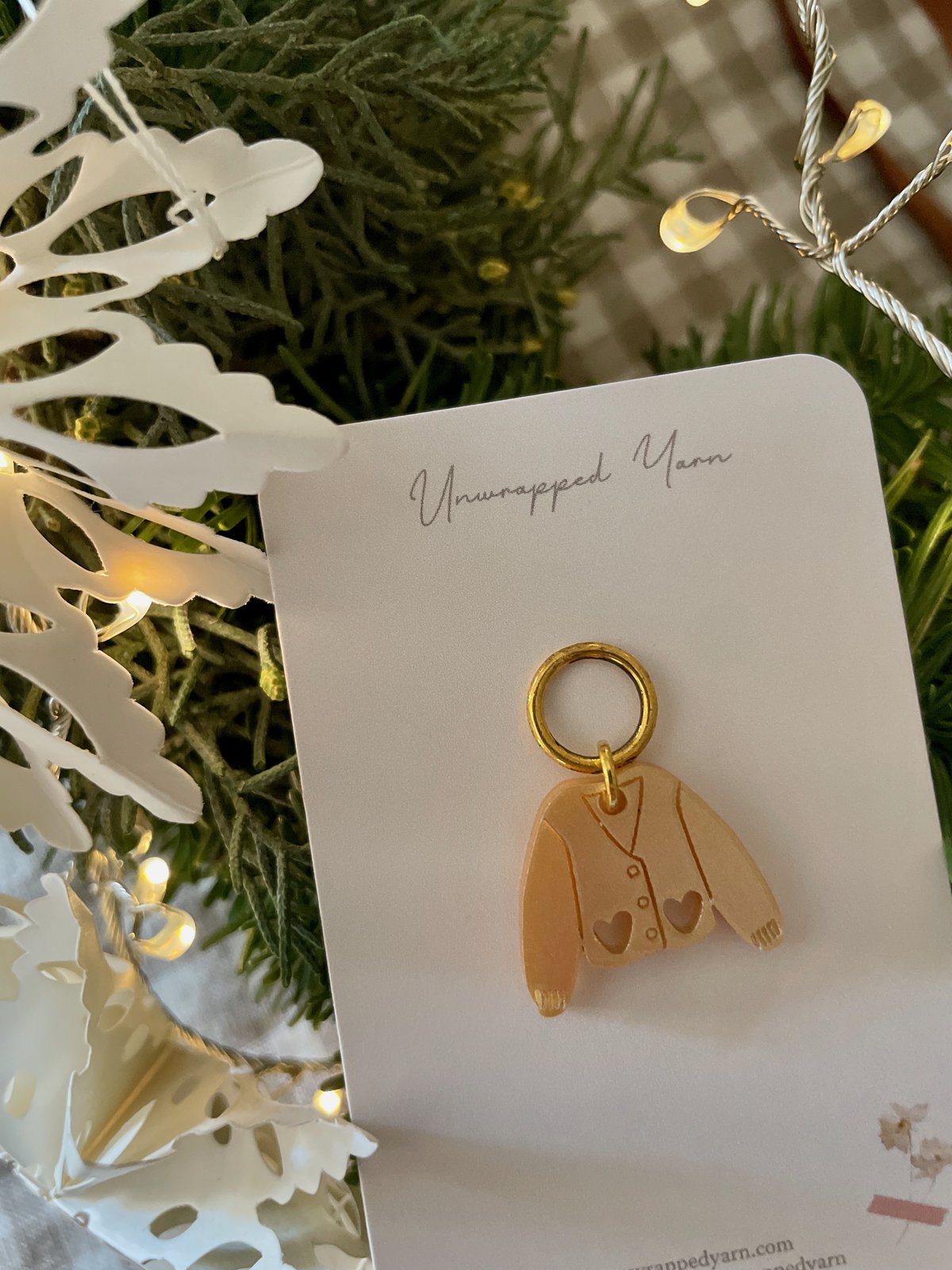 Delicate jewelry charm featuring a sweater design on a white card, surrounded by festive greenery and warm string lights.