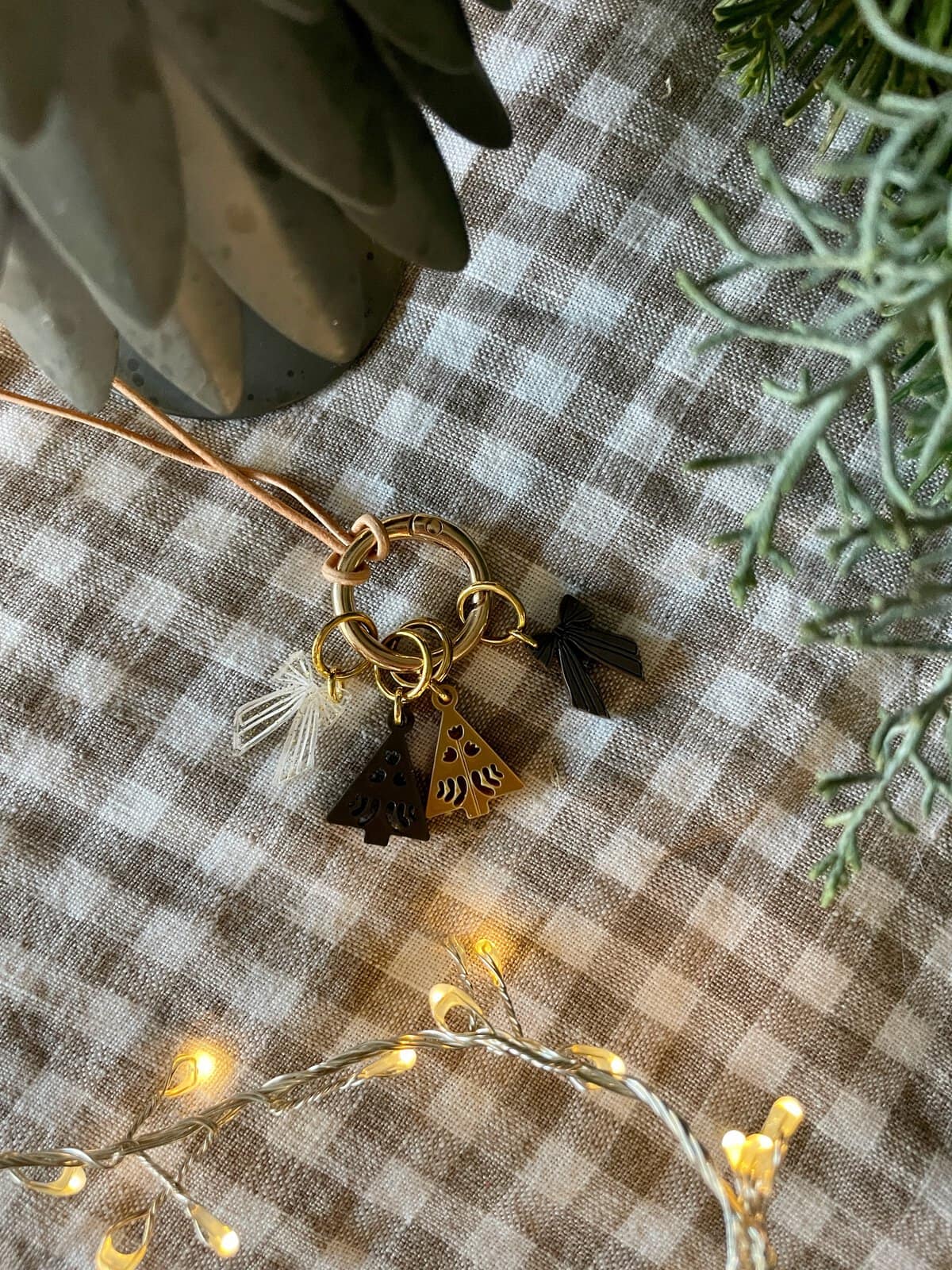Chic keychain with holiday tree charms on checkered fabric, surrounded by greenery and warm fairy lights.