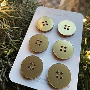 Shiny Gold Buttons on White Card with Christmas Tree and Lights in Background.
