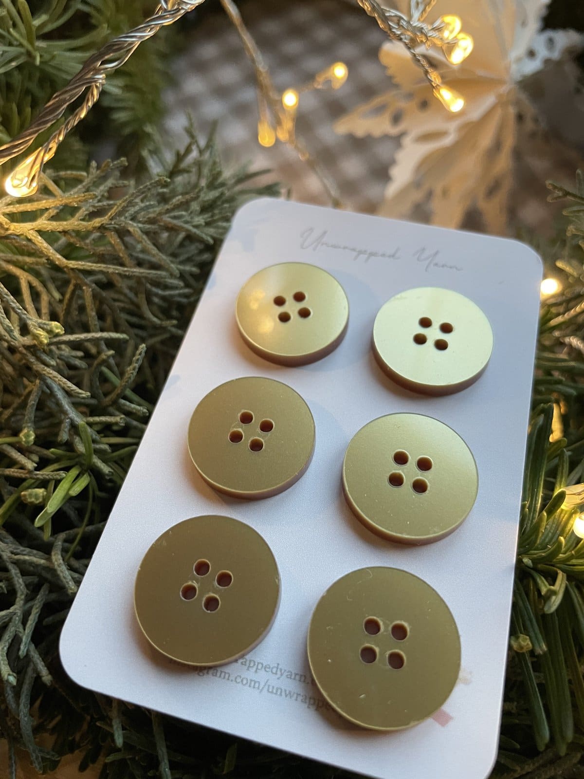 Shiny Gold Buttons on White Card with Christmas Tree and Lights in Background.