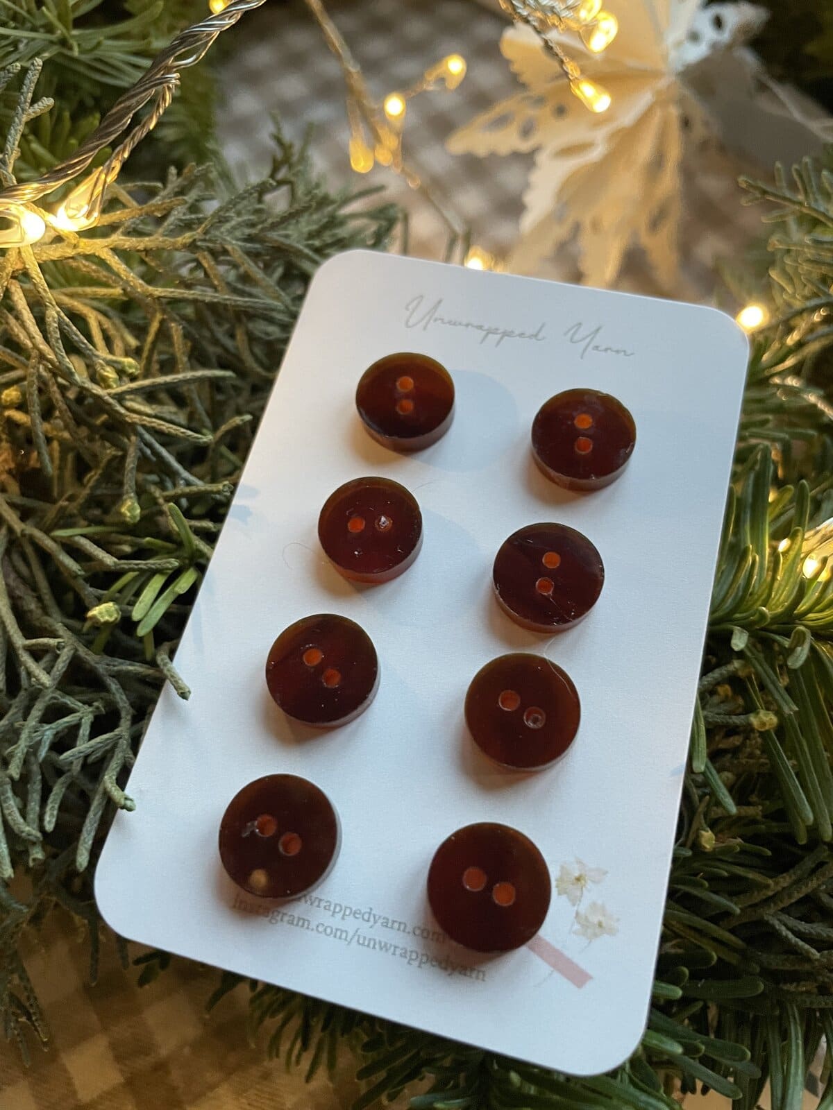Round dark brown buttons on a white card, surrounded by greenery and holiday lights. Perfect for knitting or sewing projects.