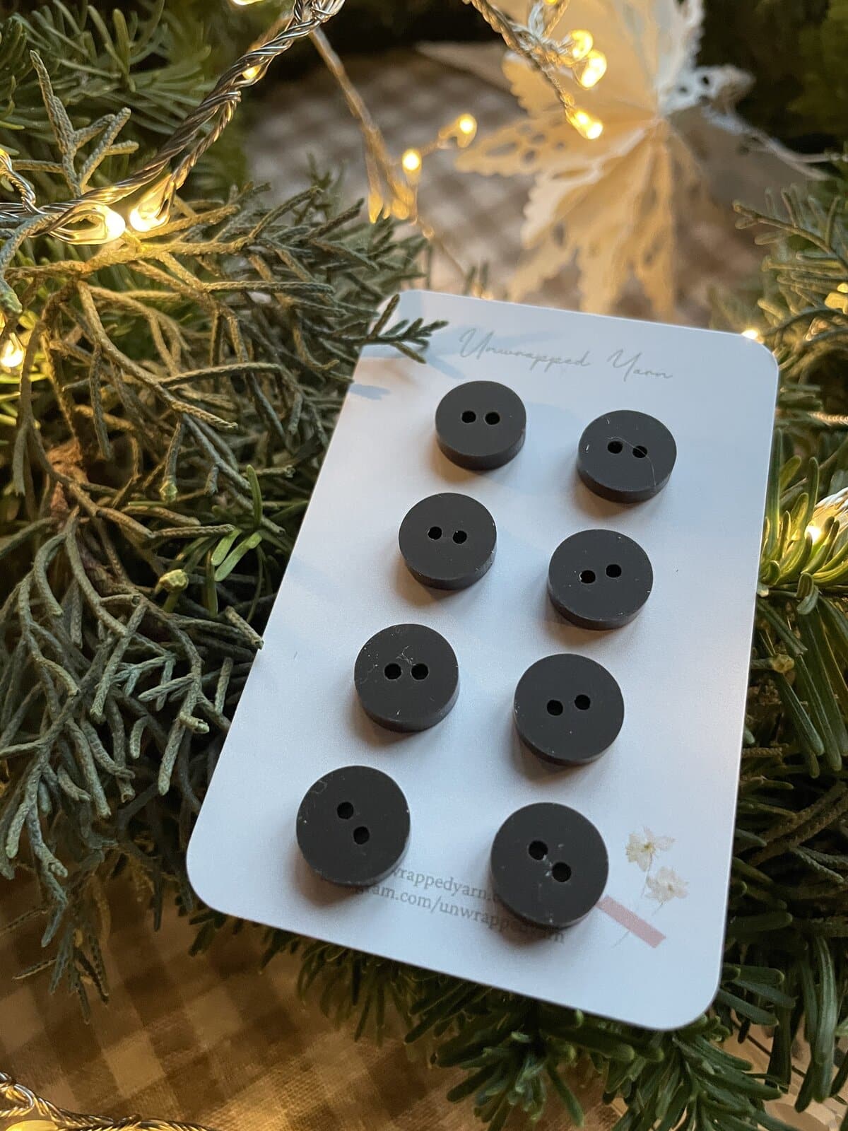 Black buttons for knitting, sewing, and crafts on a white card, surrounded by greenery and festive lights.