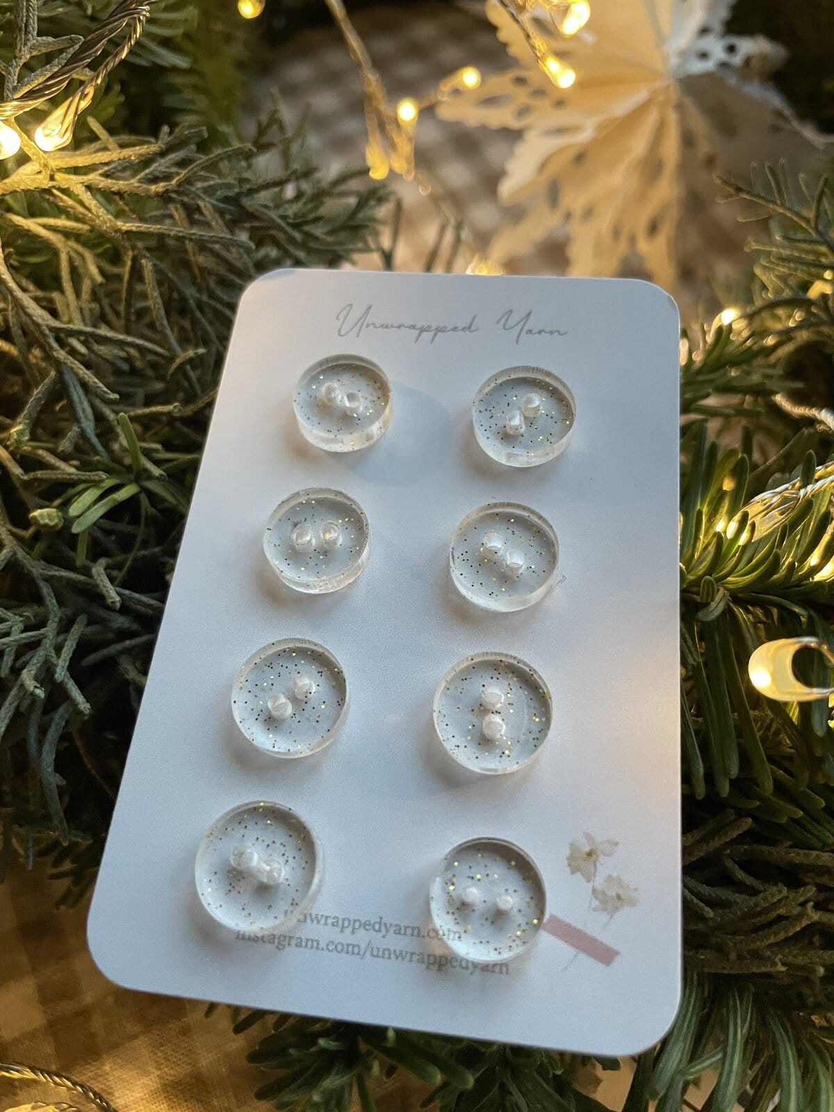 Shimmering clear resin buttons with gold glitter on a white card, surrounded by festive greenery and warm fairy lights.